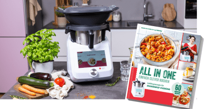 Vordergrund: Cover des All-in-One-Kochbuches; Hintergrund: Monsieur Cuisine mit dem Dampfgaraufsatz auf einer Arbeitsfläche.