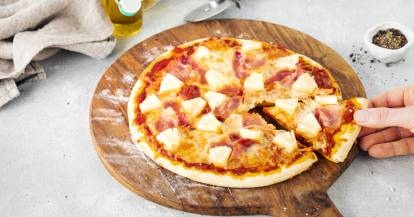 Hawaiipizza, ein Stück wird von einer Hand angehoben.