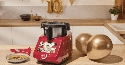 Der Monsieur Cuisine Smart in rot auf einer Holzarbeitsplatte, daneben zwei goldene Luftballons