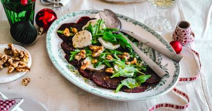 Rote-Bete-Carpaccio mit Salat und Ziegenkäse auf einer hellen Platte. Daneben ein Glas mit Weihn und Weihnachtsdeko