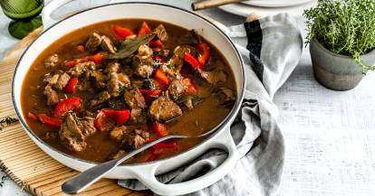 eine weiße Schale mit Griffen, die mit Gulasch gefüllt ist, daneben stehen Kräuter