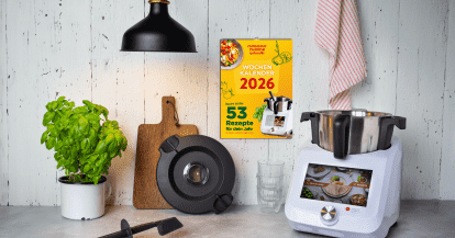 Eine Arbeitsplatte mit dem Monsieur Cuisine, Basilikum und einem Holzbrett. An der Wand hängt der ZauberMix Kalender 2026