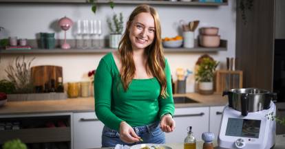 Junge Frau mit langen Haaren und grünem Pullover steht mit Monsieur Cuisine in der Küche