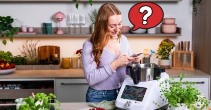 Junge Frau mit Smartphone schaut in den Monsieur Cuisine, neben ihrem Kopf ein Fragezeichen