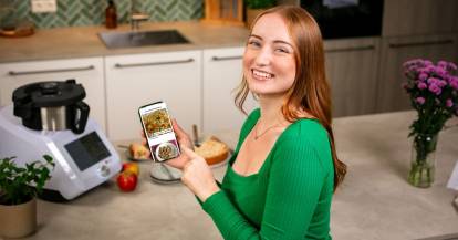 Junge Frau mit Smartphone lächelt in die Kamera, im Hintergrund ein Monsieur Cuisine