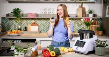 Junge Frau lacht, in der Hand hält sie ein Eisenpräparat und eine kleine Flasche mit Saft, sie steht in einer Küche
