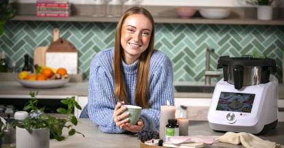 Eine junge Frau in einer Küche, lächelnd mit einem Becher in beiden Händen