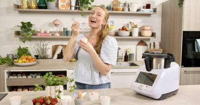 Junge Frau in einer Küche mit Monsieur Cuisine und einem Becher mit Eis in der Hand