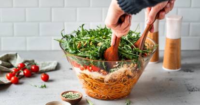 Zwei Hände mit Holzlöffeln nehmen Salat aus einer Schüssel, die Spaghetti, Parmesan, Tomaten und Rucola enthält