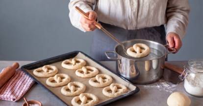 Jemand hält eine Kelle mit einer Brezel darauf und legt sie in einen Topf mit Wasser, daneben ein Blech mit Brezeln, ungebacken