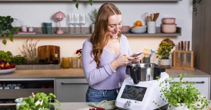 Junge Frau schaut auf ihr Handy, sie steht in einer Küche neben dem Monsieur Cuisine