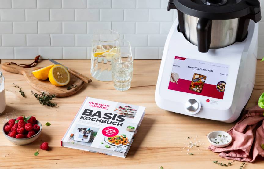 Das Basis-Kochbuch liegt auf einer Arbeitsfläche neben dem Monsieur Cuisine und einigen Zutaten.