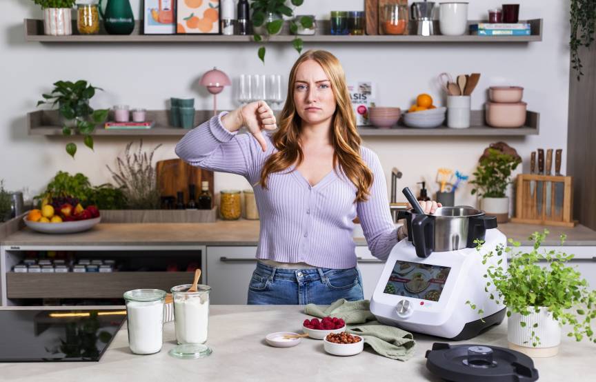 Frau steht in einer Küche neben dem Monsieur Cuisine und hält den Daumen runter