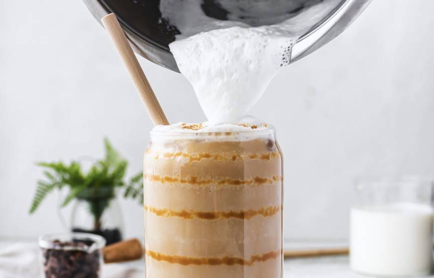 Jemand gießt Milchschaum aus dem Mixbehälter des Monsieur Cuisine auf einen Kaffee