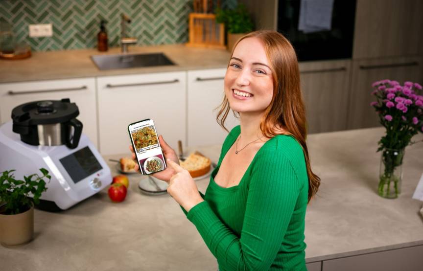 Junge Frau mit Smartphone lächelt in die Kamera, im Hintergrund ein Monsieur Cuisine