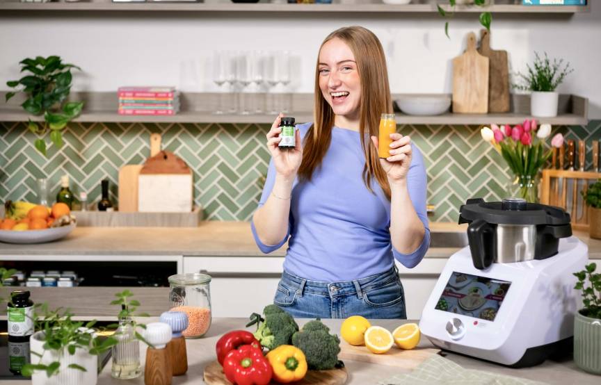 Junge Frau lacht, in der Hand hält sie ein Eisenpräparat und eine kleine Flasche mit Saft, sie steht in einer Küche