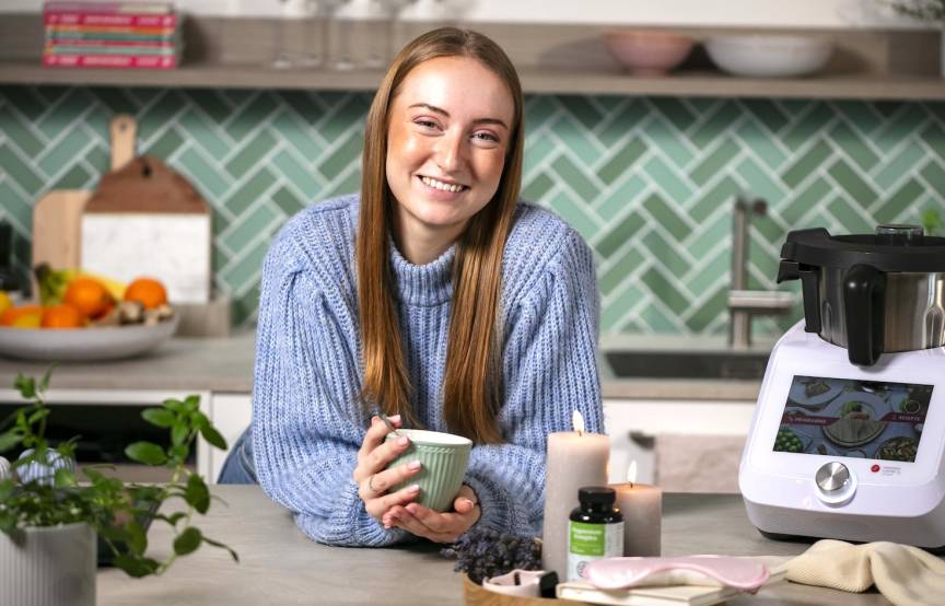 Eine junge Frau in einer Küche, lächelnd mit einem Becher in beiden Händen