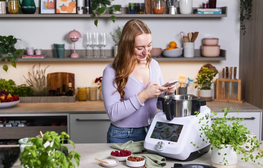 Junge Frau mit Smartphone in einer Küche, vor ihr der Monsieur Cuisine
