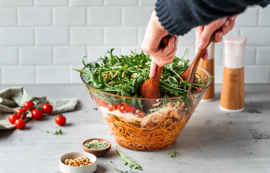 Zwei Hände mit Holzlöffeln nehmen Salat aus einer Schüssel, die Spaghetti, Parmesan, Tomaten und Rucola enthält