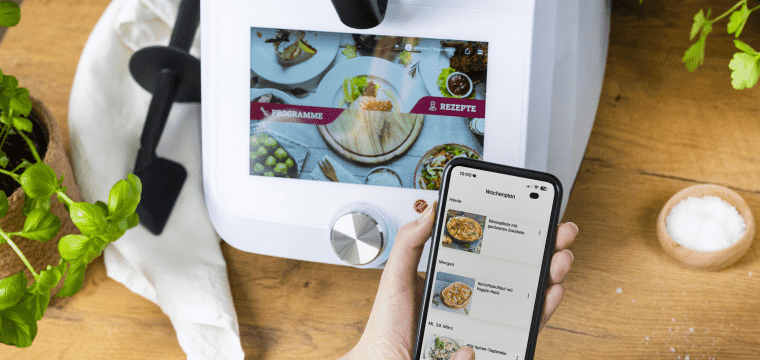 Monsieur Cuisine im Hintergrund, im Vordergrund eine Hand, die ein Handy hält. Auf dem Display ist die ZauberMix App mit dem Wochenplaner.