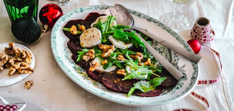 Rote-Bete-Carpaccio mit Salat und Ziegenkäse auf einer hellen Platte. Daneben ein Glas mit Weihn und Weihnachtsdeko