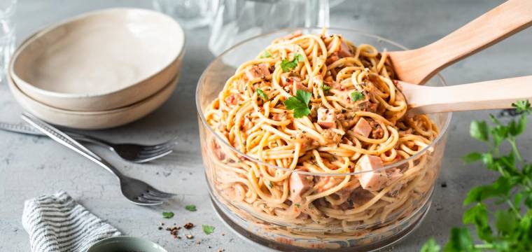 Eine Glasschale mit Spaghettisalat auf grauer Arbeitsplatte, daneben Teller und Kräuter