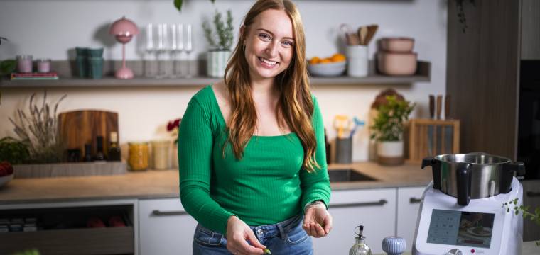 Junge Frau mit langen Haaren und grünem Pullover steht mit Monsieur Cuisine in der Küche