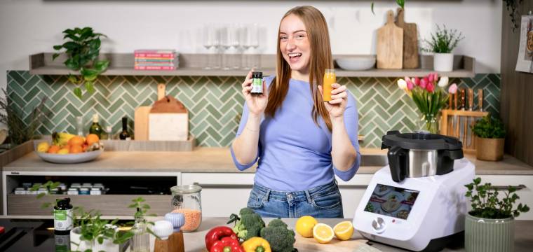 Junge Frau lacht, in der Hand hält sie ein Eisenpräparat und eine kleine Flasche mit Saft, sie steht in einer Küche