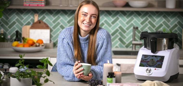 Eine junge Frau in einer Küche, lächelnd mit einem Becher in beiden Händen Eine junge Frau in einer Küche, lächelnd mit einem Becher in beiden Händen