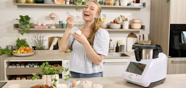 Junge Frau in einer Küche mit Monsieur Cuisine und einem Becher mit Eis in der Hand