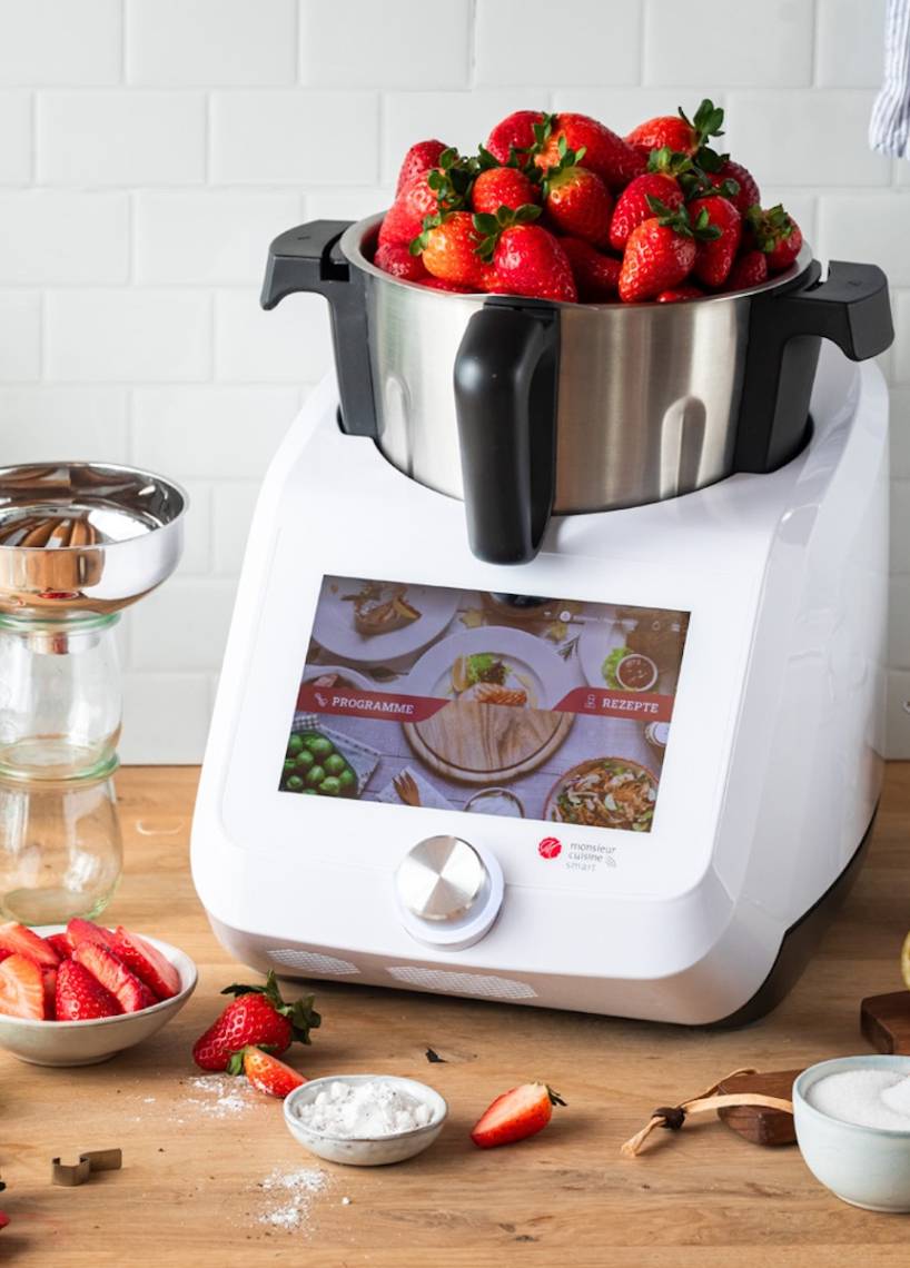 Der Monsieur Cuisine auf einer Arbeitsplatte aus Holz, im Mixbehälter sind Erdbeeren, die über den Rand schauen Der Monsieur Cuisine auf einer Arbeitsplatte aus Holz, im Mixbehälter sind Erdbeeren, die über den Rand schauen