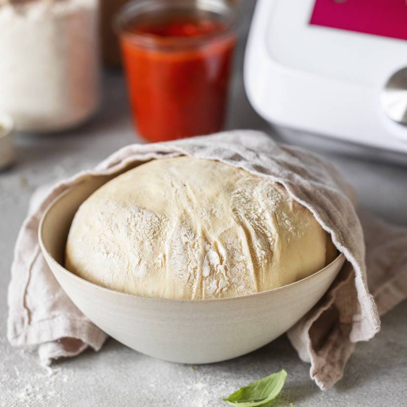 Aufgegangener Pizzateig in einer Schüssel, halb abgedeckt mit einem Küchentuch.