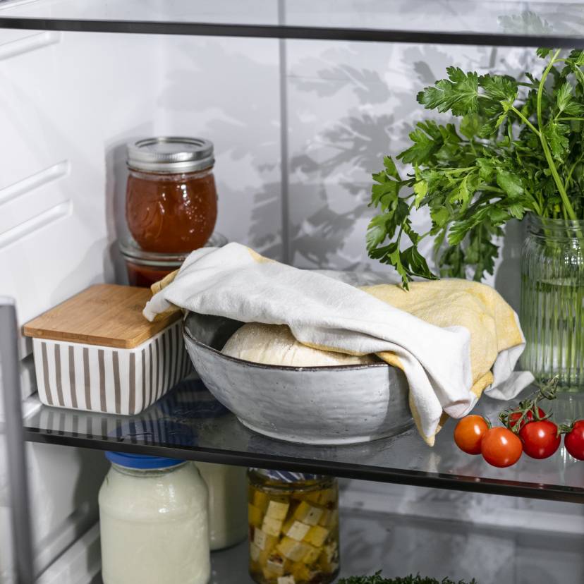 Abgedeckter Pizzateig in einem gefüllten Kühlschrank.