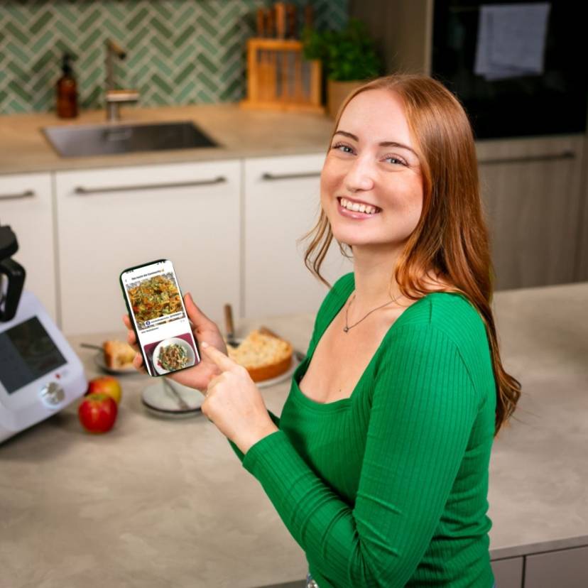 Junge Frau mit Smartphone lächelt in die Kamera, im Hintergrund ein Monsieur Cuisine