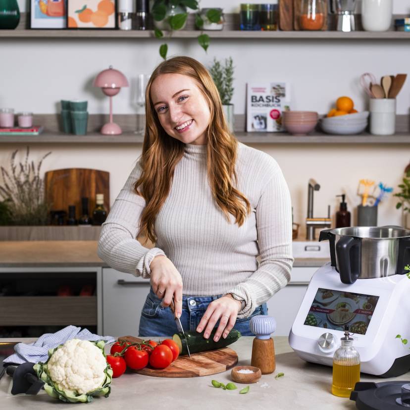 Junge Frau mit langen Haaren schneidet Gemüse, neben ihr steht ein Monsieur Cuisine